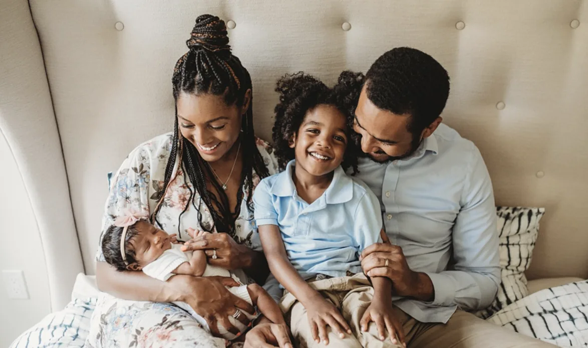A family of four sitting together on a bed, parents smiling down at their newborn baby and young son
